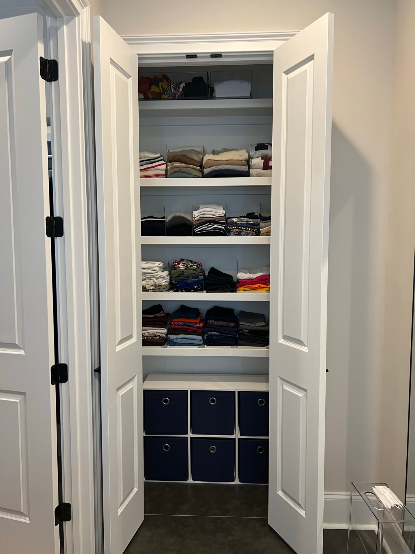 Neatly organized white reach-in closet with folded clothing, acrylic dividers, and navy storage bins